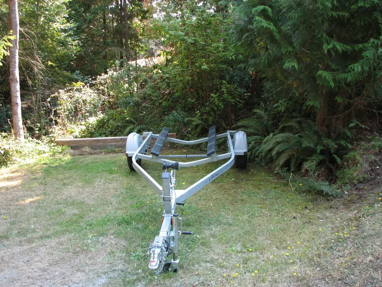Empty single-axle boat trailer parked on grass, with its hitch, winch and hull supports visible.